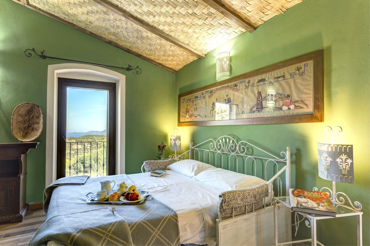 A bedroom with a bed, a bedside table, a lamp, a painting, and a view of the outdoors.