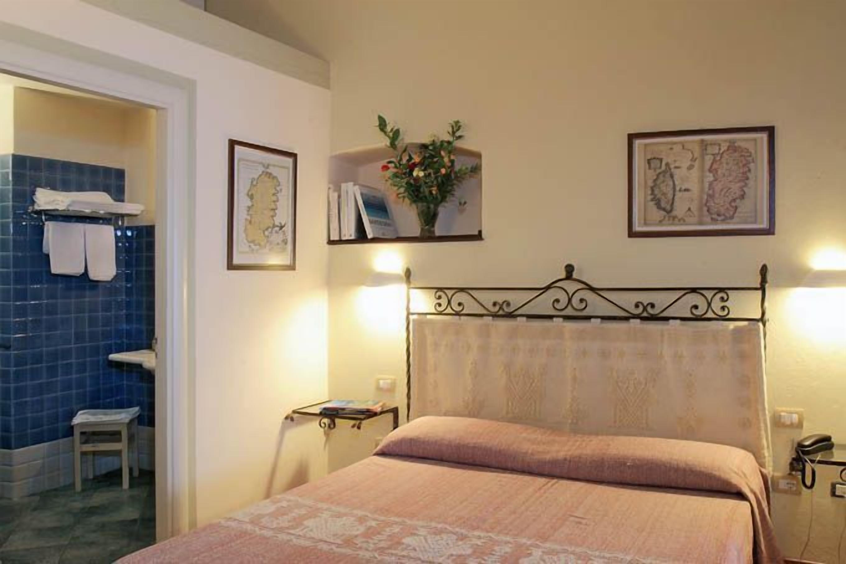 Ein Hotelzimmer mit Bett, einem Badezimmer mit blauen Fliesen, einem kleinen Tisch mit einem Buch und gerahmten Bildern an der Wand.