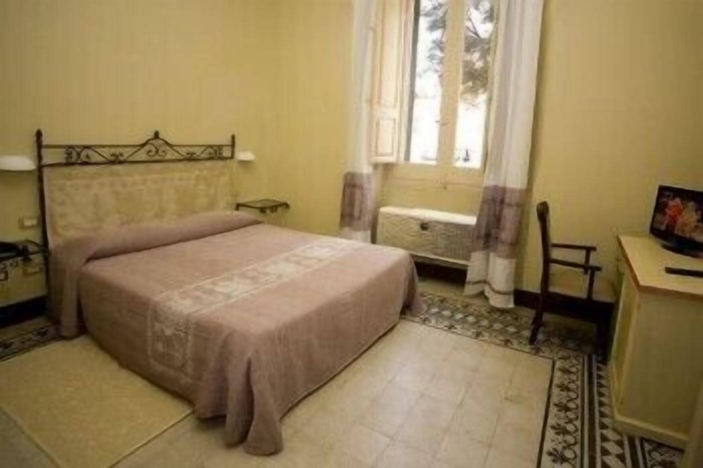 A bedroom with a bed, a bedside table, a chair, a window with curtains, and a small desk with a picture frame.