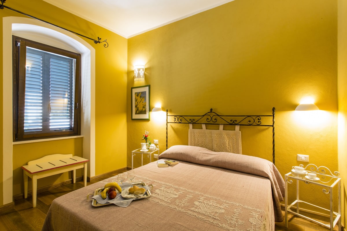 A hotel room with a bed, bedside table, lamp, and a window with blinds.