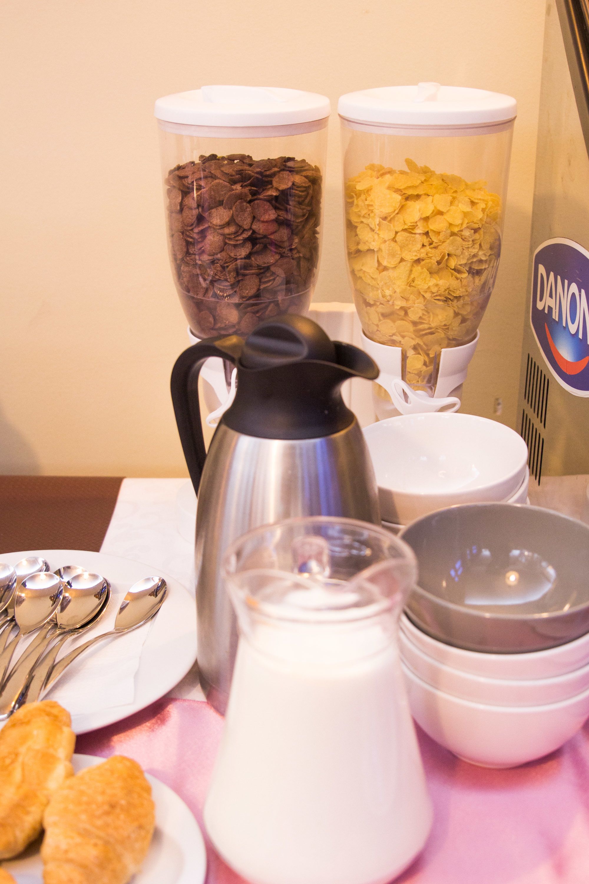 A breakfast buffet setup with containers of chocolate and cornflakes, a milk pitcher, and a coffee pot.