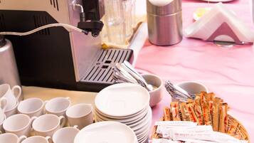 Una mesa de buffet con una cafetera, diversos alimentos y cubiertos sobre un mantel rosa.