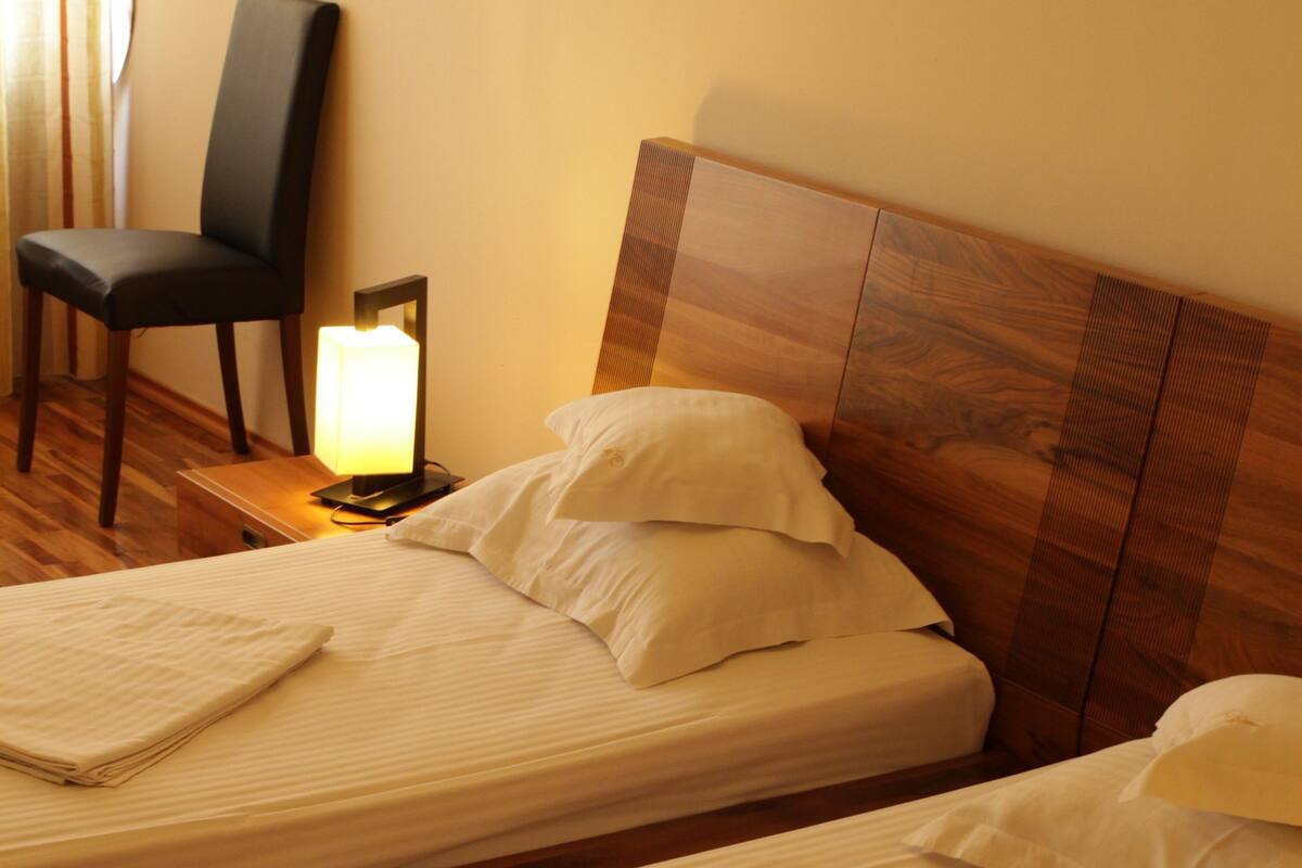 A hotel room with two single beds, a nightstand with a lamp, a chair, and a wooden headboard.