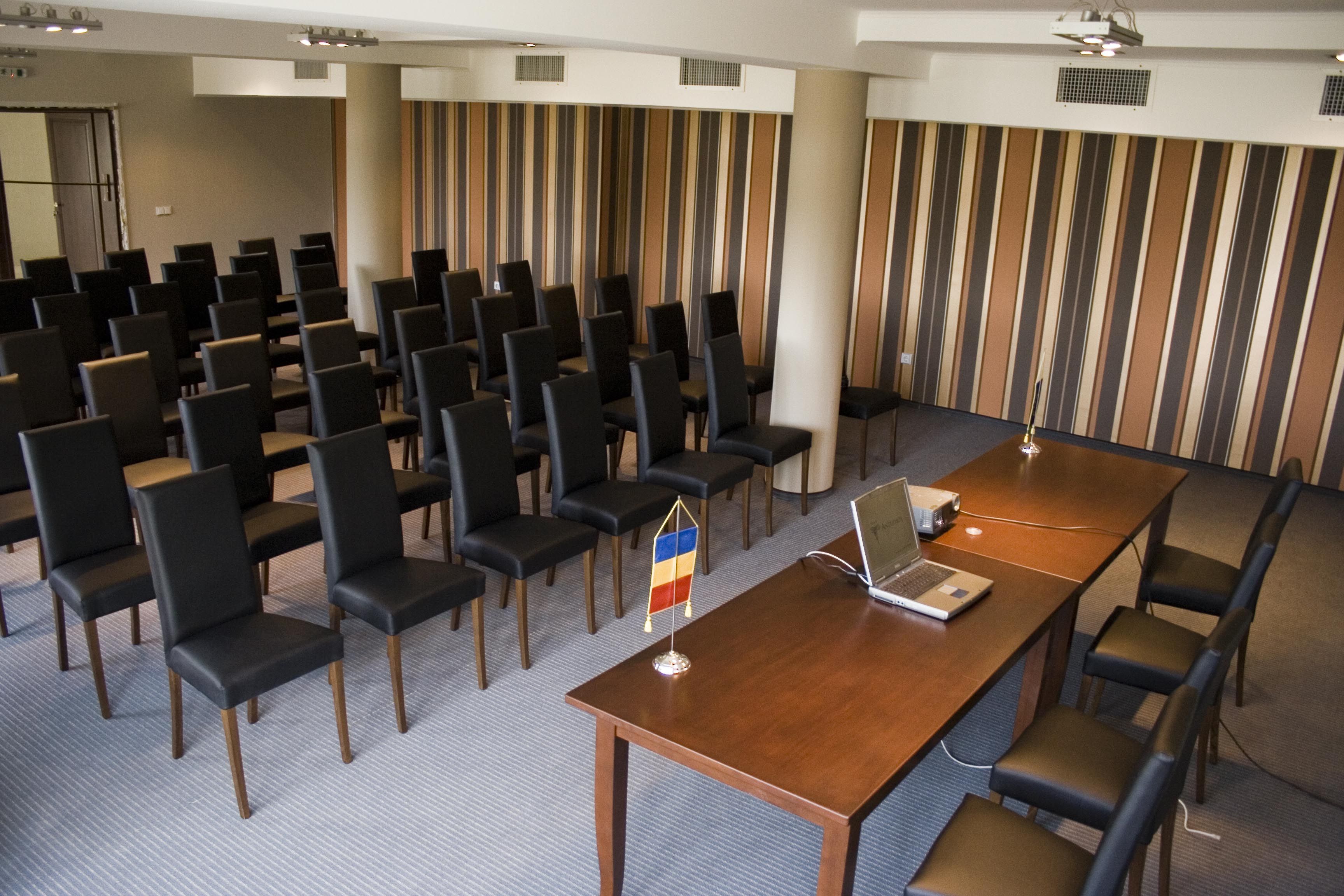 A conference room with rows of black chairs, a wooden table with a laptop, and a flag on the table.