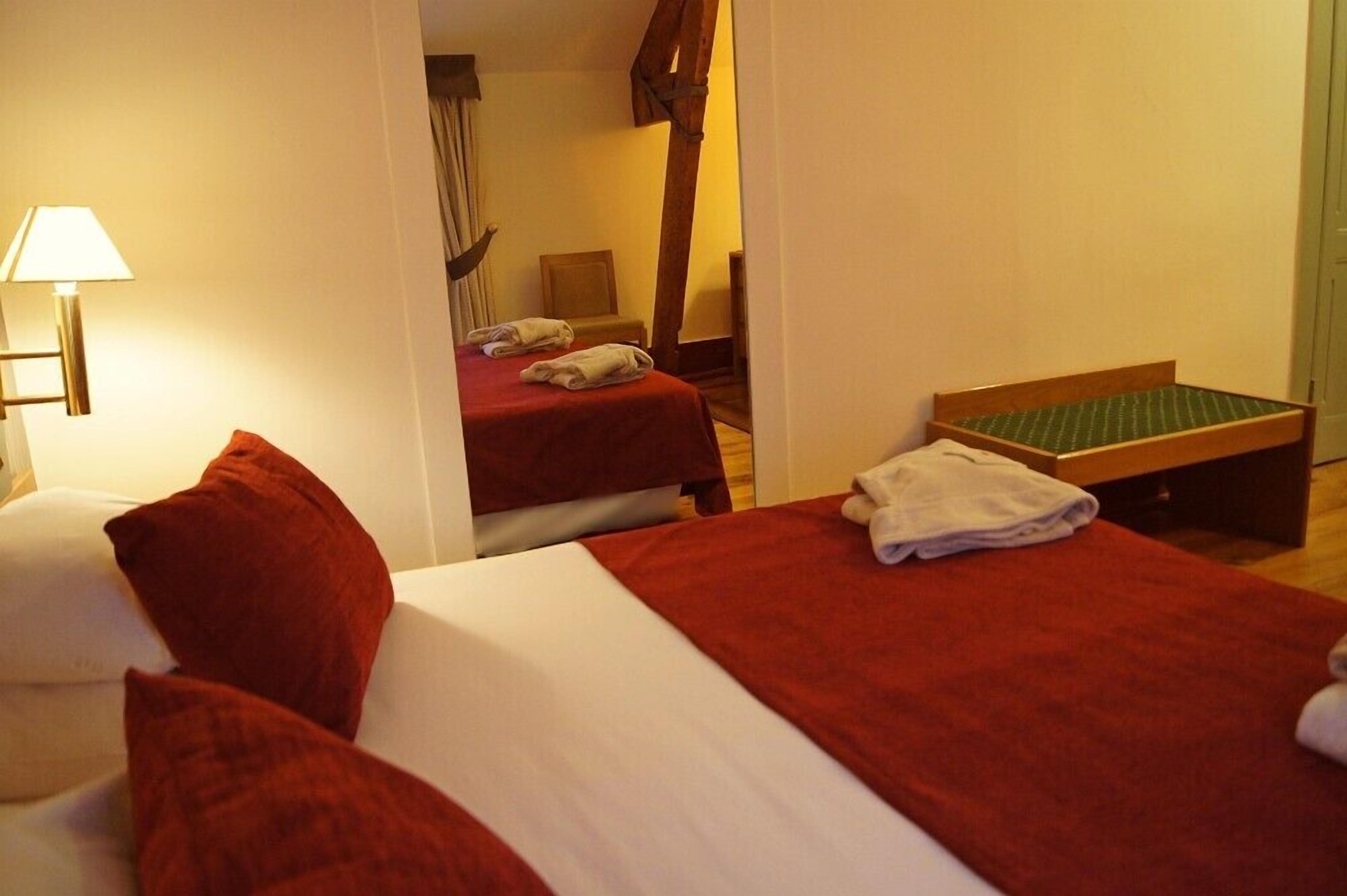 Ein Hotelzimmer mit zwei Betten, eines mit roter Bettdecke und das andere mit weißer Bettdecke. Auf einer hölzernen Bank mit grüner Sitzfläche liegt ein weißes Handtuch. Eine Lampe ist auf der linken Seite des Bildes zu sehen.
