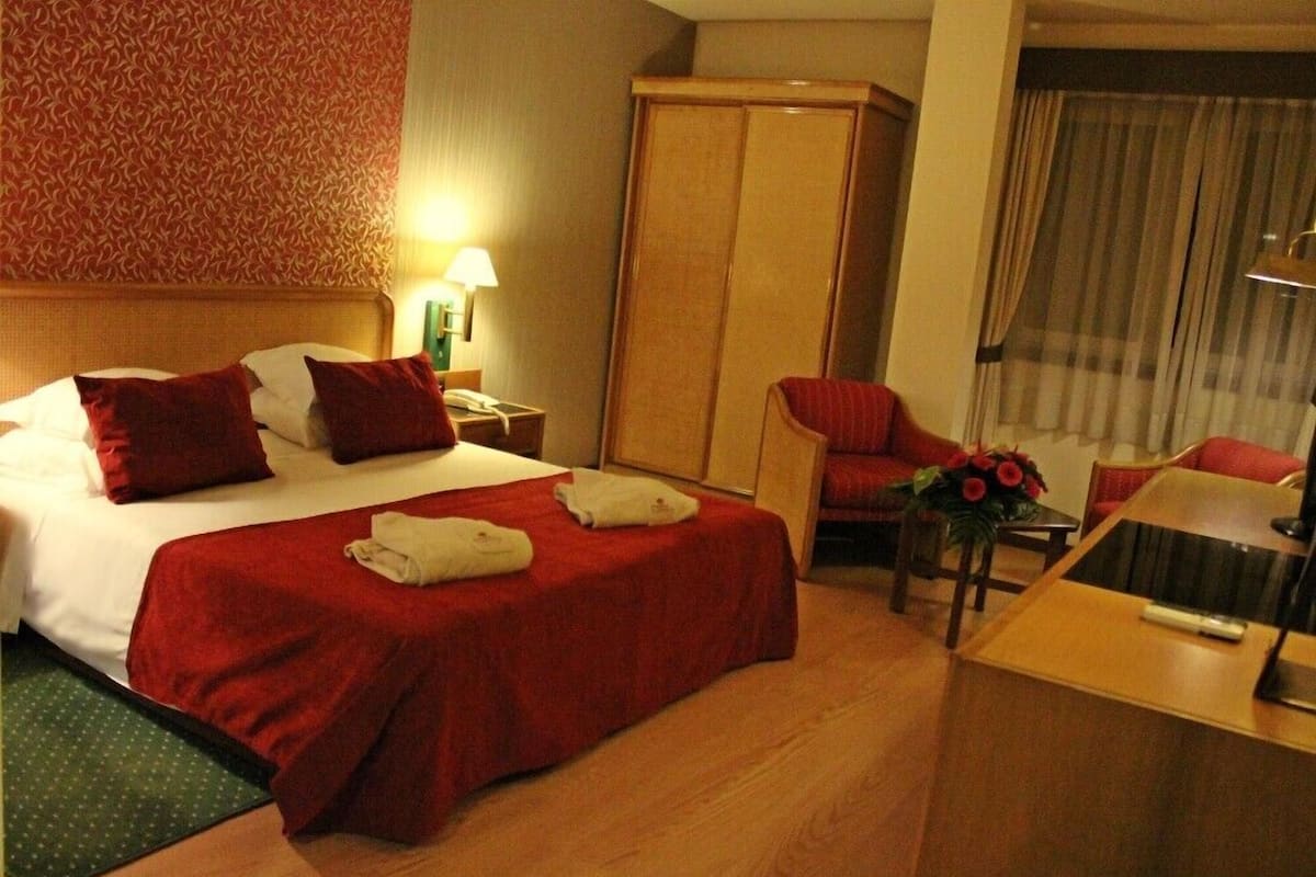 Une chambre d’hôtel comprenant un lit, un bureau, une chaise, une armoire et une fenêtre avec des rideaux.