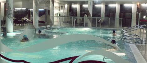 Une piscine intérieure dotée de plusieurs couloirs, d’un espace détente avec des chaises et d’une grande fresque murale.