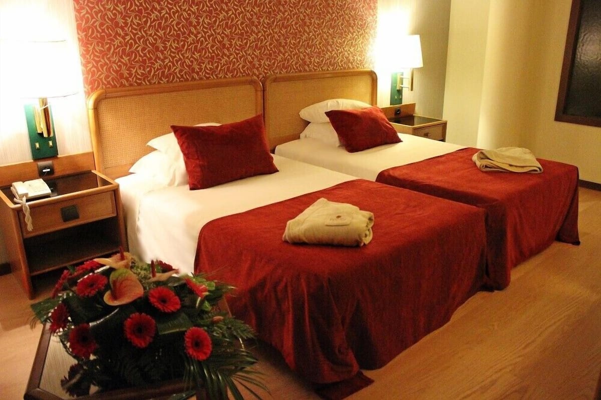 Une chambre d’hôtel avec deux lits, une literie rouge, une table de chevet avec une lampe et un arrangement floral.
