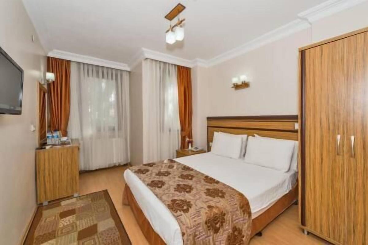 A hotel room with a bed, a wardrobe, a desk with a chair, a television, and a window with curtains.