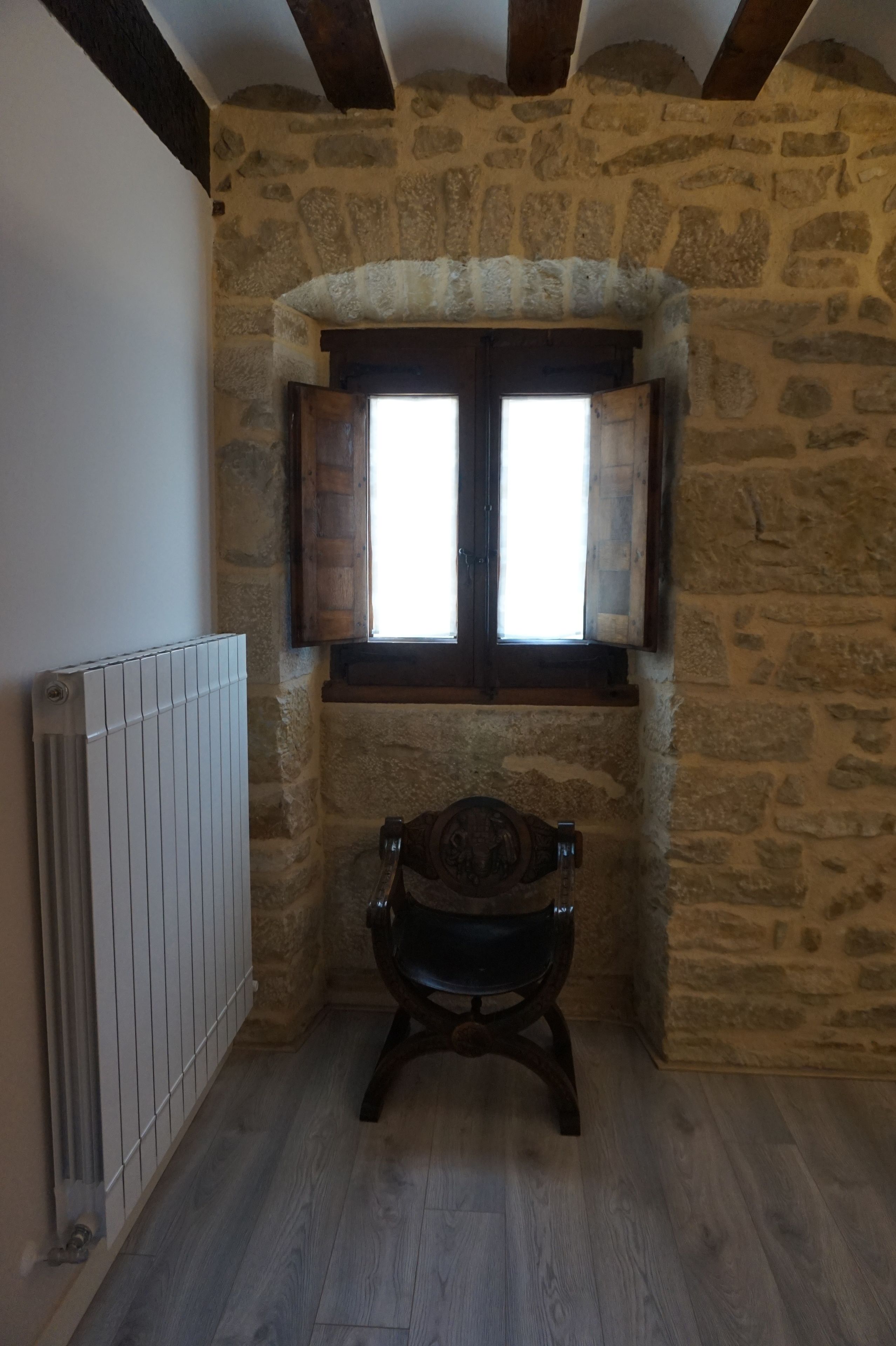 Una habitación con paredes de piedra, una silla de madera, un radiador y una ventana con contraventanas de madera.