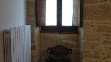 Una habitación con paredes de piedra, una silla de madera, un radiador y una ventana con contraventanas de madera.