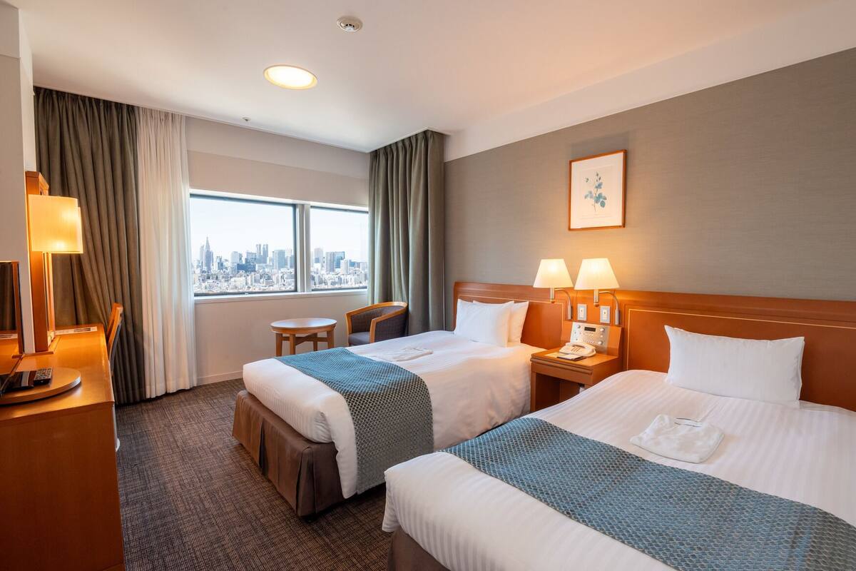 A hotel room with two beds, a desk with a lamp, a chair, a nightstand with a phone, and a window with a city view.