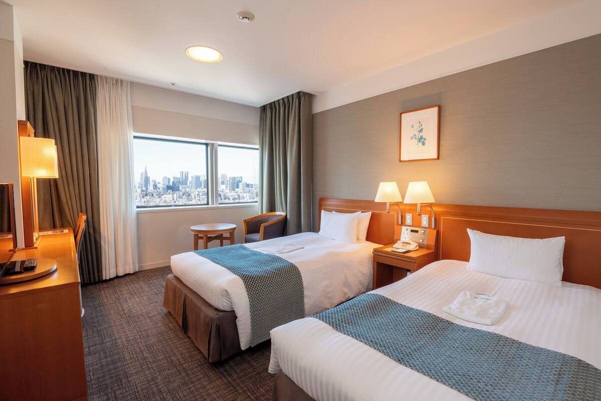 A hotel room with two beds, a desk, a chair, a window with a city view, and a painting on the wall.
