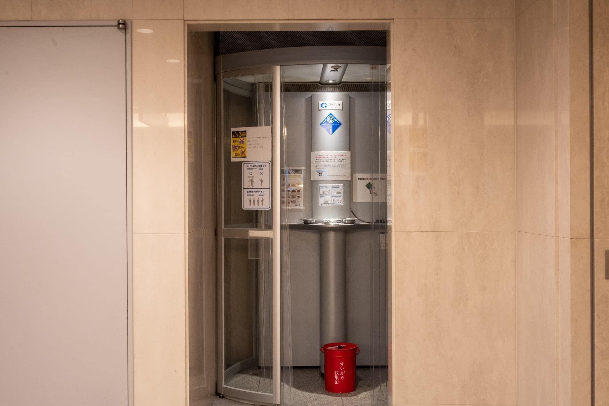 A glass elevator with a red bucket at its base, a sign with emergency instructions, and a logo on the wall.