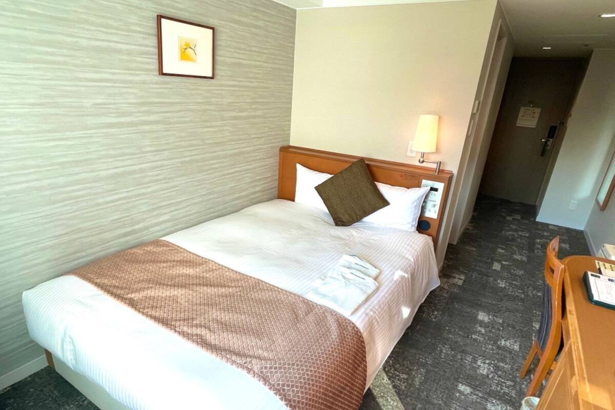 A hotel room with a bed, a bedside table, a lamp, a framed picture on the wall, and a door leading to another room.