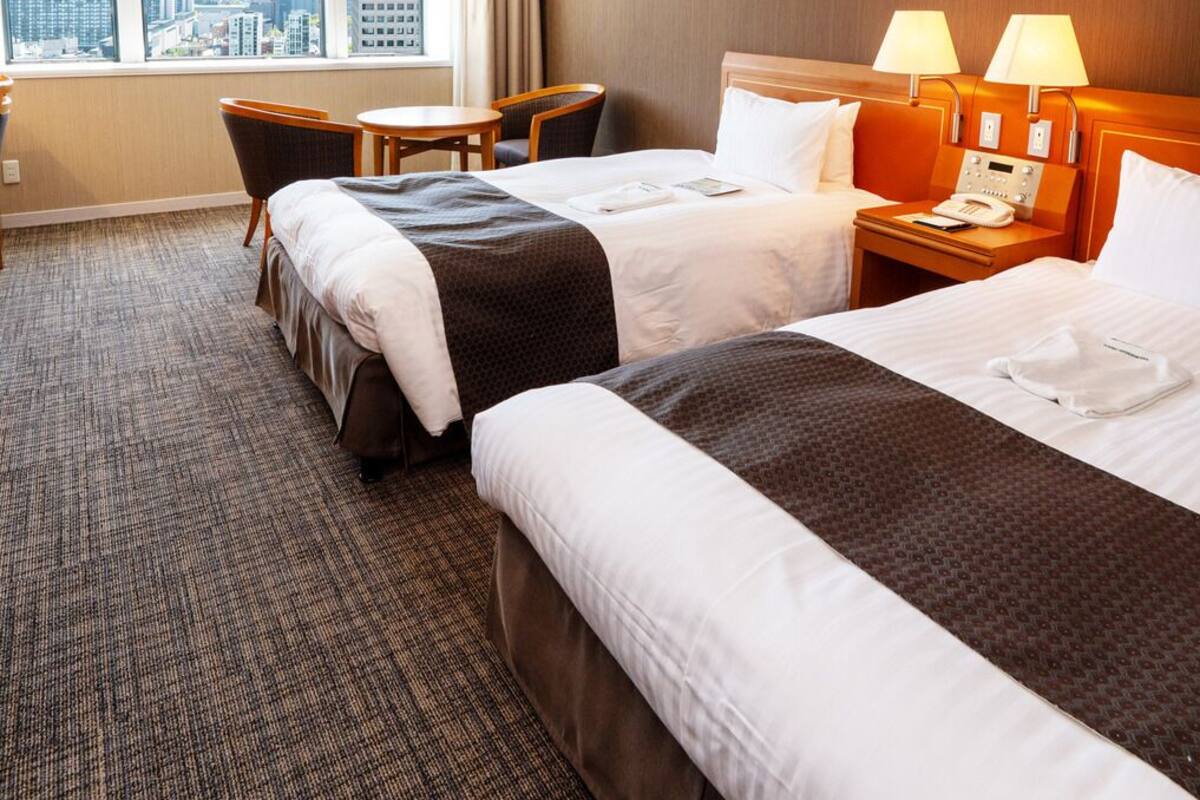 A hotel room with two beds, a desk, a chair, and a large window with a city view.