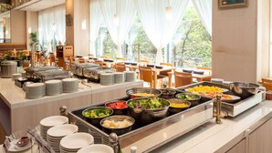 A buffet setup with various food items, a counter with stacked plates, and a view of the outdoors through large windows. - Toshi Center Hotel Tokyo (Tokyo)