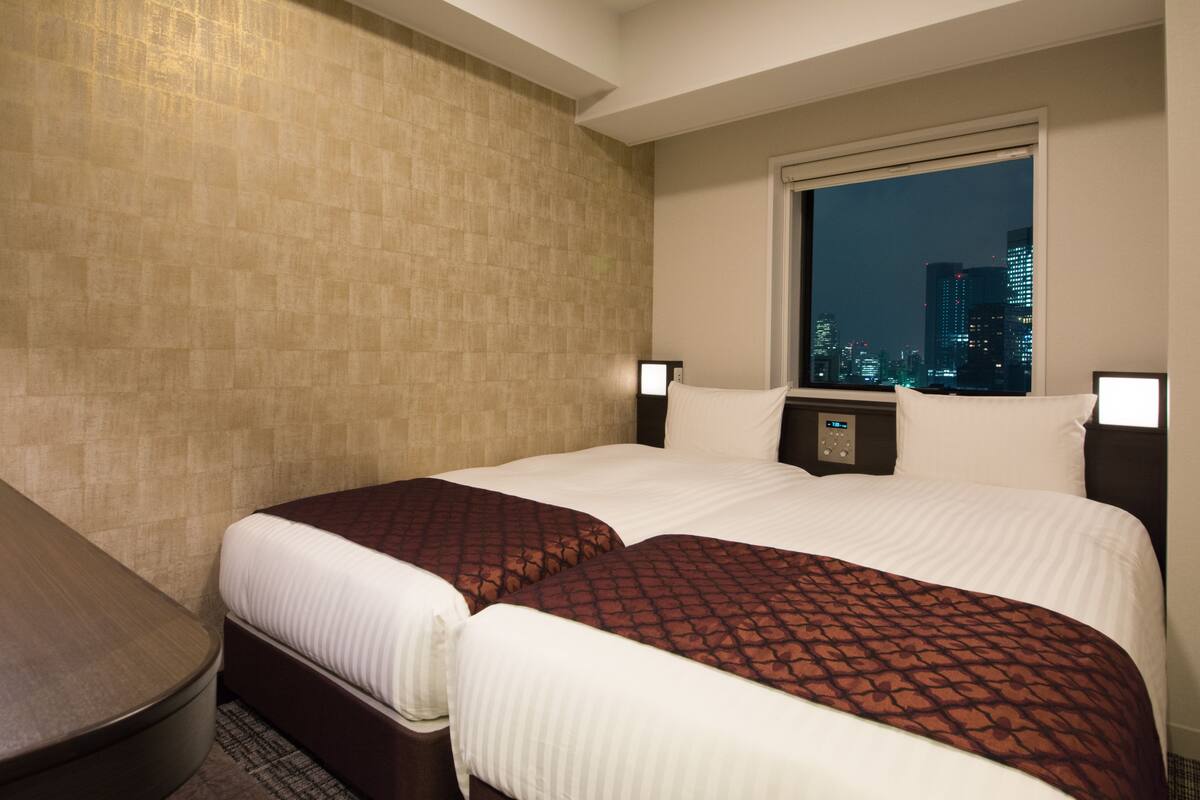 A hotel room with two beds, a city view through a large window, a bedside table, and a wall-mounted alarm clock.