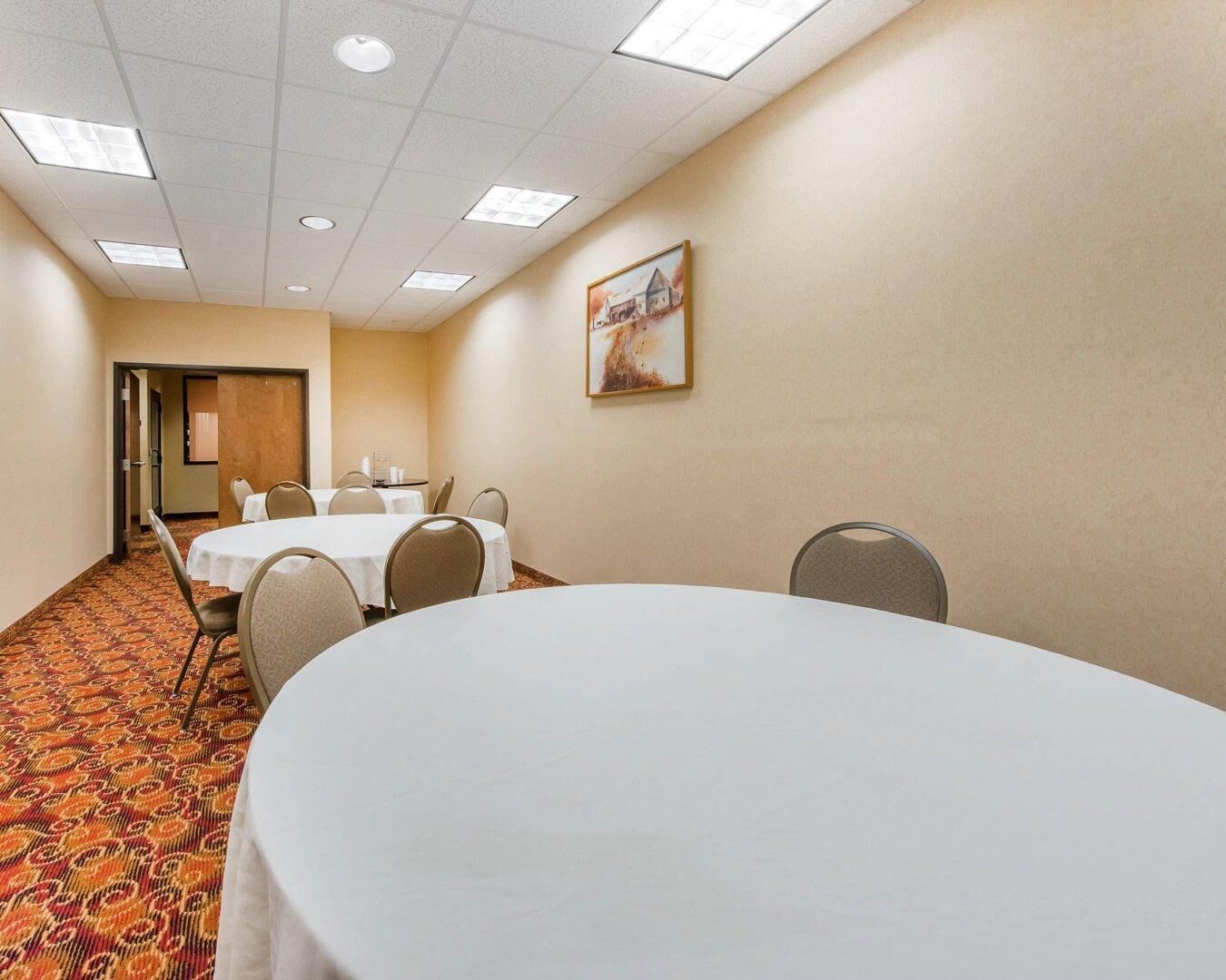Une salle de conférence dotée d’une grande table ovale, de petites tables rondes et de chaises, ainsi que d’un tapis à motifs et d’un tableau accroché au mur.