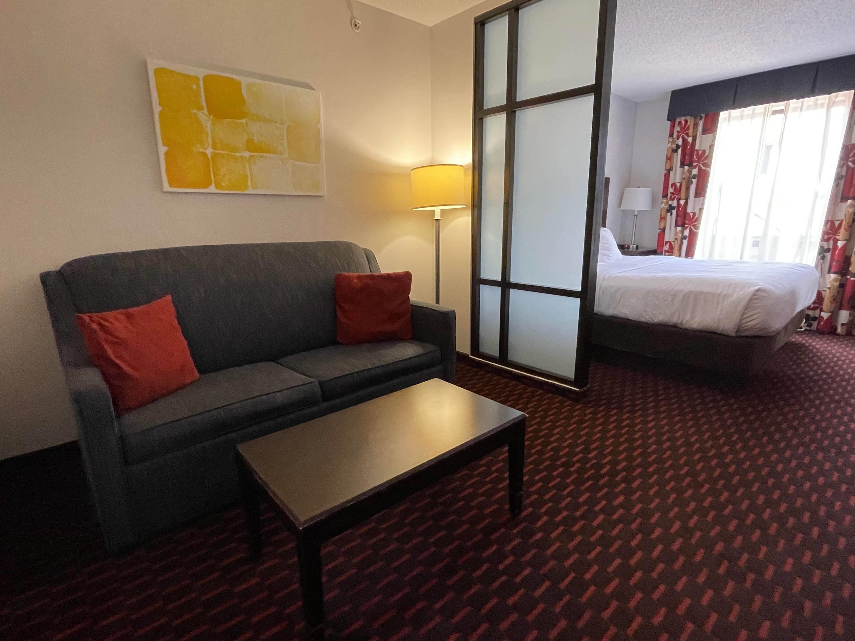 A hotel room with a grey sofa, red cushions, a small table, a bed with white bedding, and a large window with curtains.