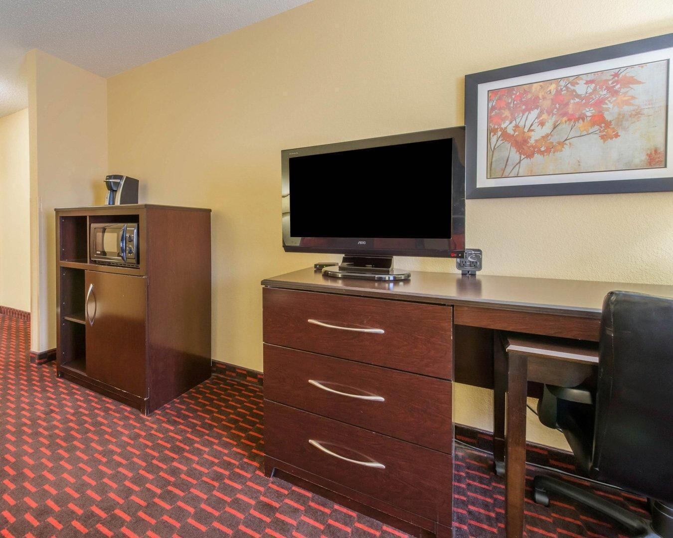 Ett hotellrum med ett skrivbord, en tv, en minibar och en inramad tavla på väggen.