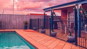 A swimming pool with a metal ladder, surrounded by a fence, and a covered seating area with chairs and potted plants.