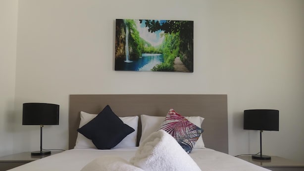 A neatly made bed with white linens and a blue bedspread, flanked by two black lamps on nightstands, and a painting of a landscape above the bed.