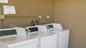 A row of white commercial dryers with control panels, a clothes line sign, and a bulletin board on a beige wall.