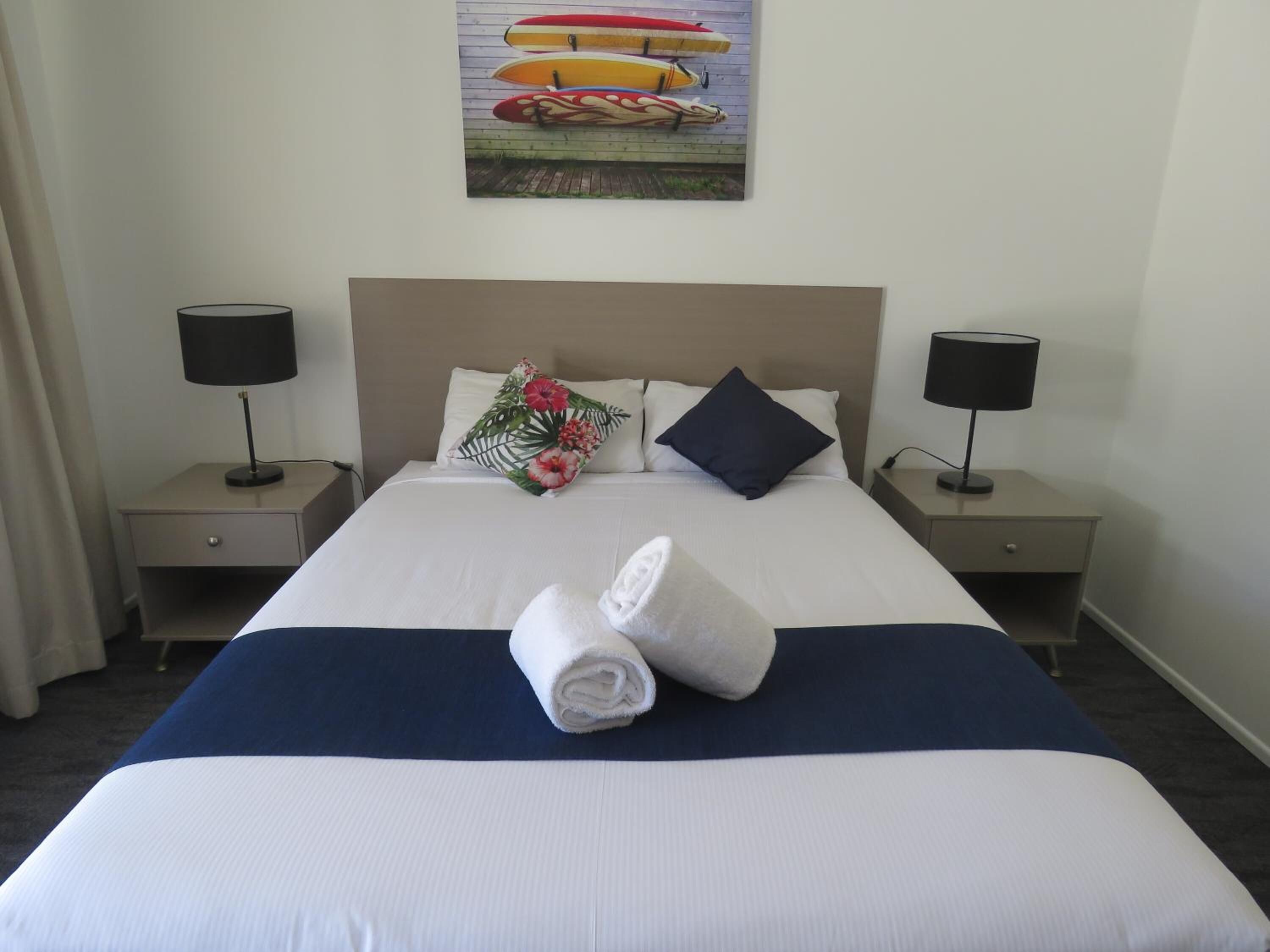 A hotel room with a neatly made bed, two bedside tables with lamps, a tropical-themed pillow, and a painting of kayaks on the wall.