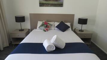A hotel room with a neatly made bed, two bedside tables with lamps, a tropical-themed pillow, and a painting of kayaks on the wall.