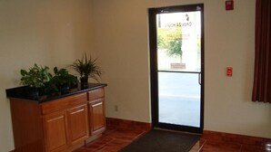 A reception area with a wooden cabinet, potted plants, a door leading outside, and a red emergency exit sign. - Winnquest Inn Near Ft. Sam Houston (San Antonio)
