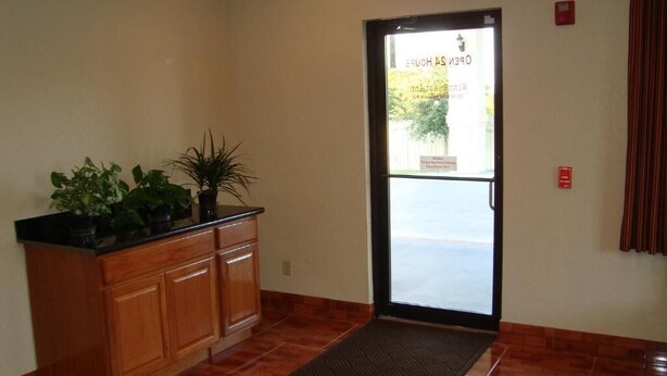 A reception area with a wooden cabinet, potted plants, a door leading outside, and a red emergency exit sign. - Winnquest Inn Near Ft. Sam Houston (San Antonio)