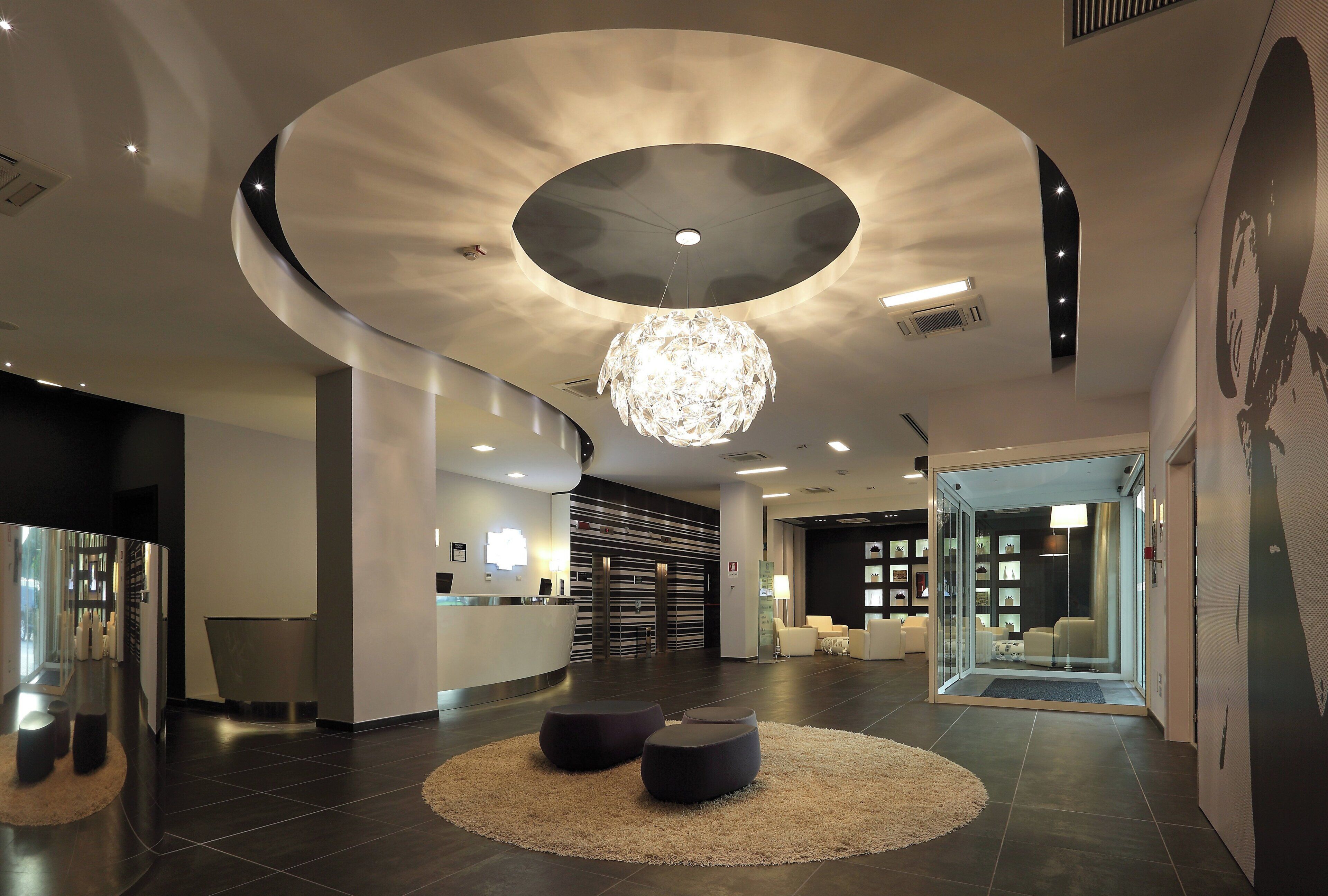 Una hall moderna con un lampadario centrale, una reception e un'area salotto con pouf rotondi.