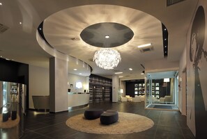 A modern lobby with a central chandelier, a reception desk, and a seating area with round ottomans. - Holiday Inn Milan Nord Zara by IHG (Cinisello Balsamo)