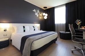 Quarto de hotel moderno com uma cama grande, uma escrivaninha com cadeira, uma mesa redonda com um vaso de flores e uma luminária fixada na parede.
