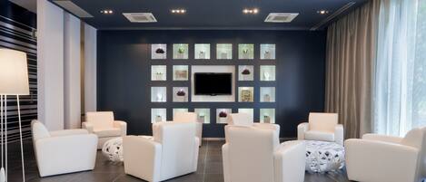 A modern lounge area with a flat-screen TV mounted on a dark wall, surrounded by white armchairs and a floor lamp.