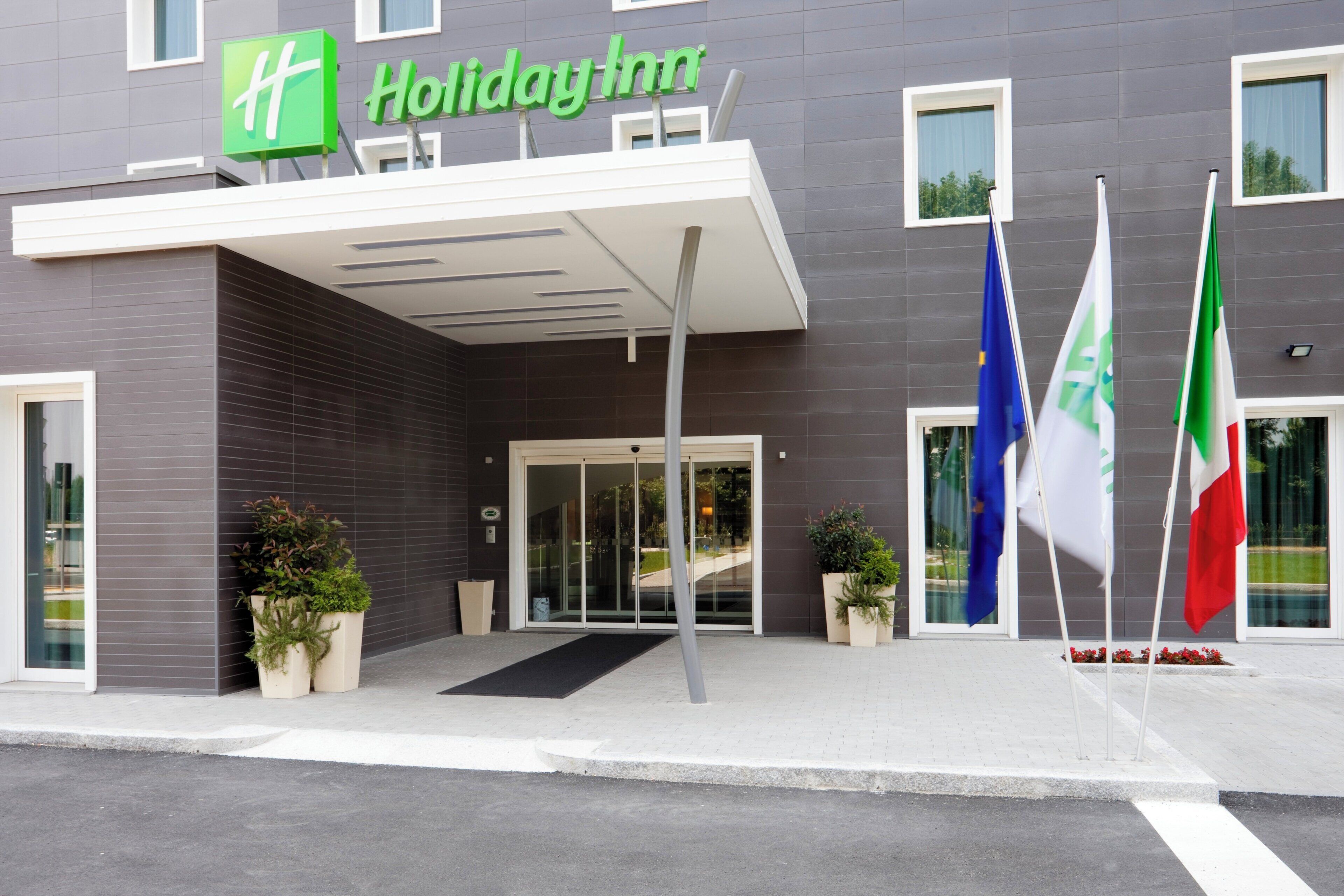 A Holiday Inn hotel entrance with a modern design, featuring a glass door entrance, potted plants, and flags.
