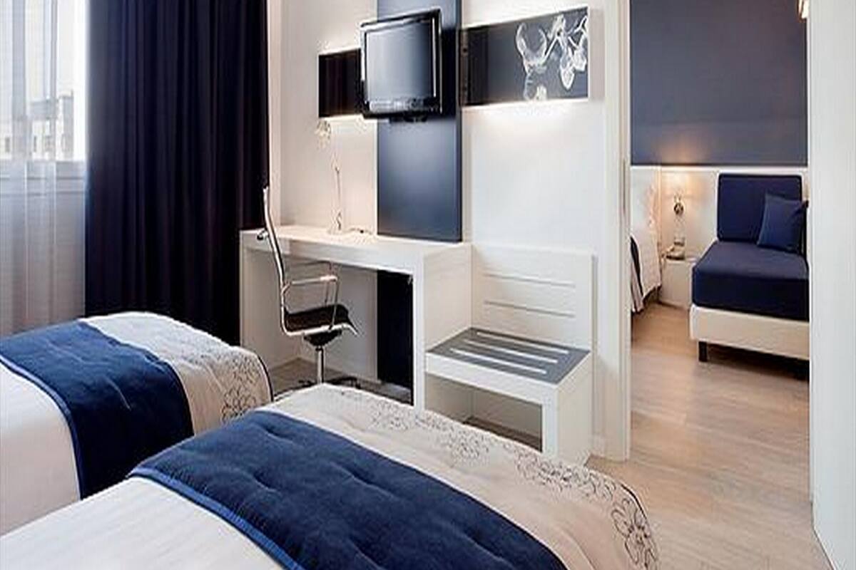 A modern hotel room with two beds, a desk with a chair, a flat-screen TV mounted on the wall, and a large window with curtains.