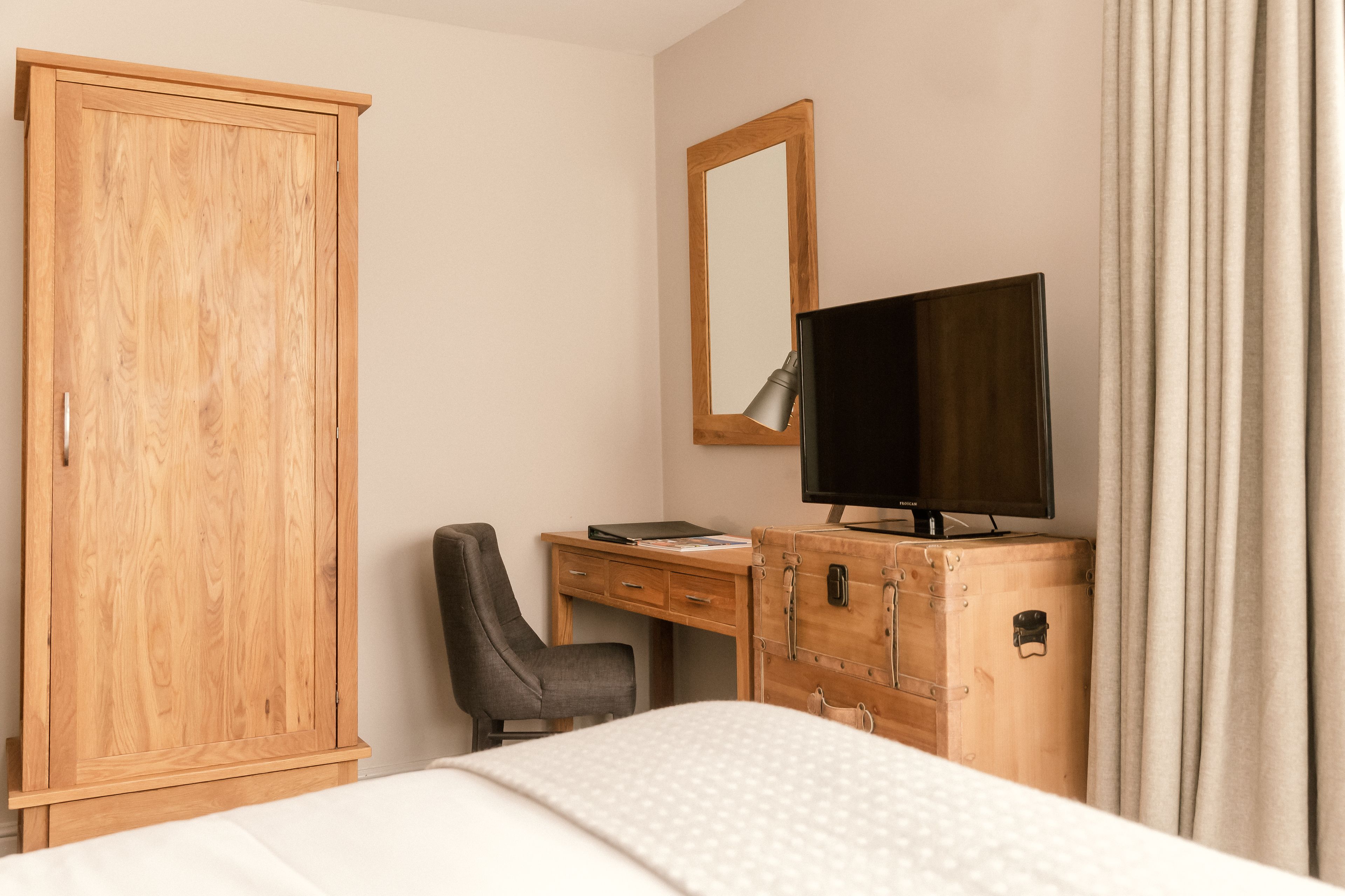 A bedroom with a wooden wardrobe, a desk with a chair, a television, and a bed.