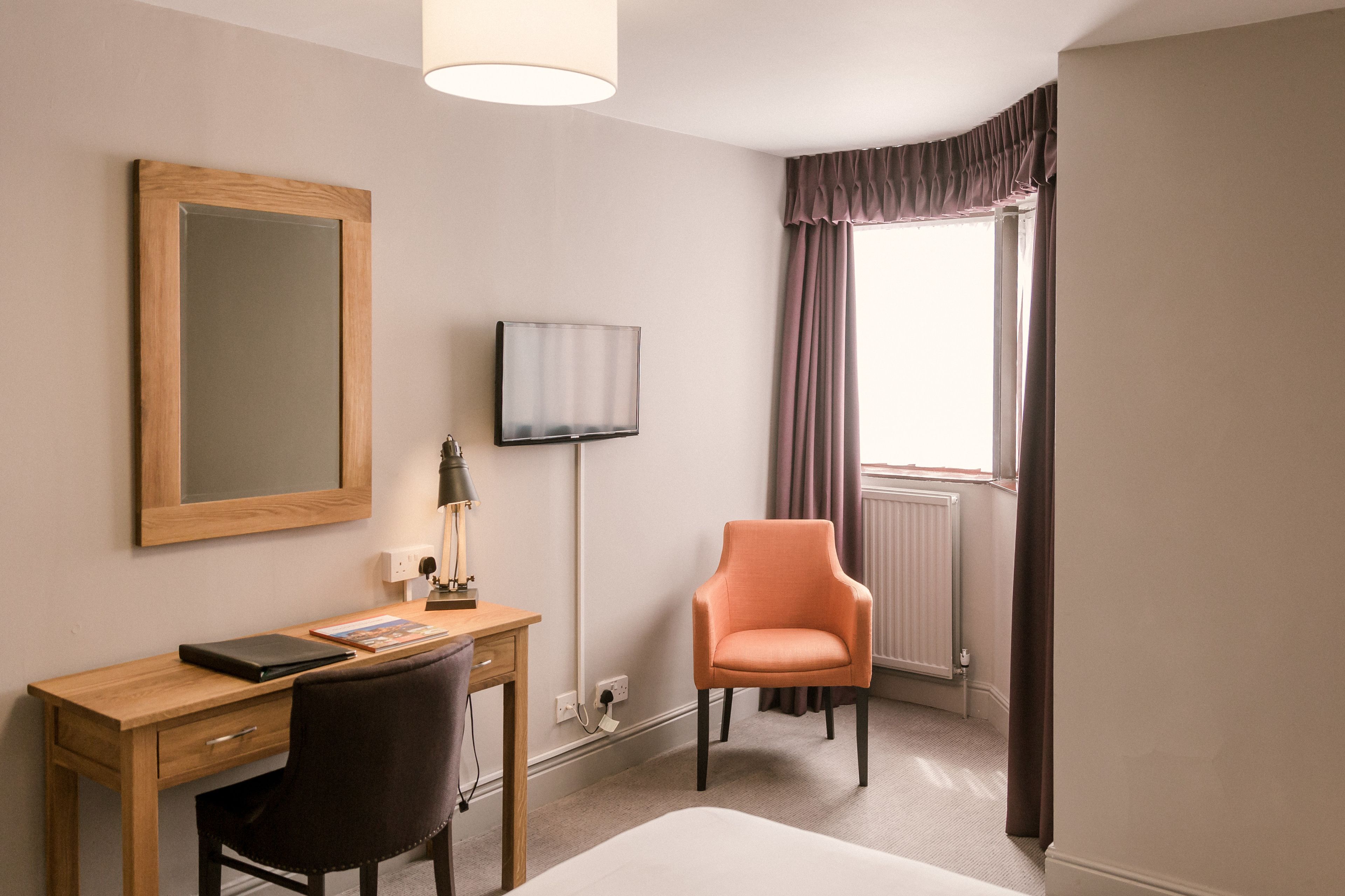 Habitación de hotel con un escritorio de madera, una silla, una cama, un televisor y un ventanal con cortinas.