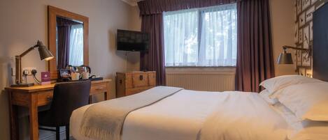 A hotel room with a bed, a desk with a computer, a chair, a TV, and a window with curtains.