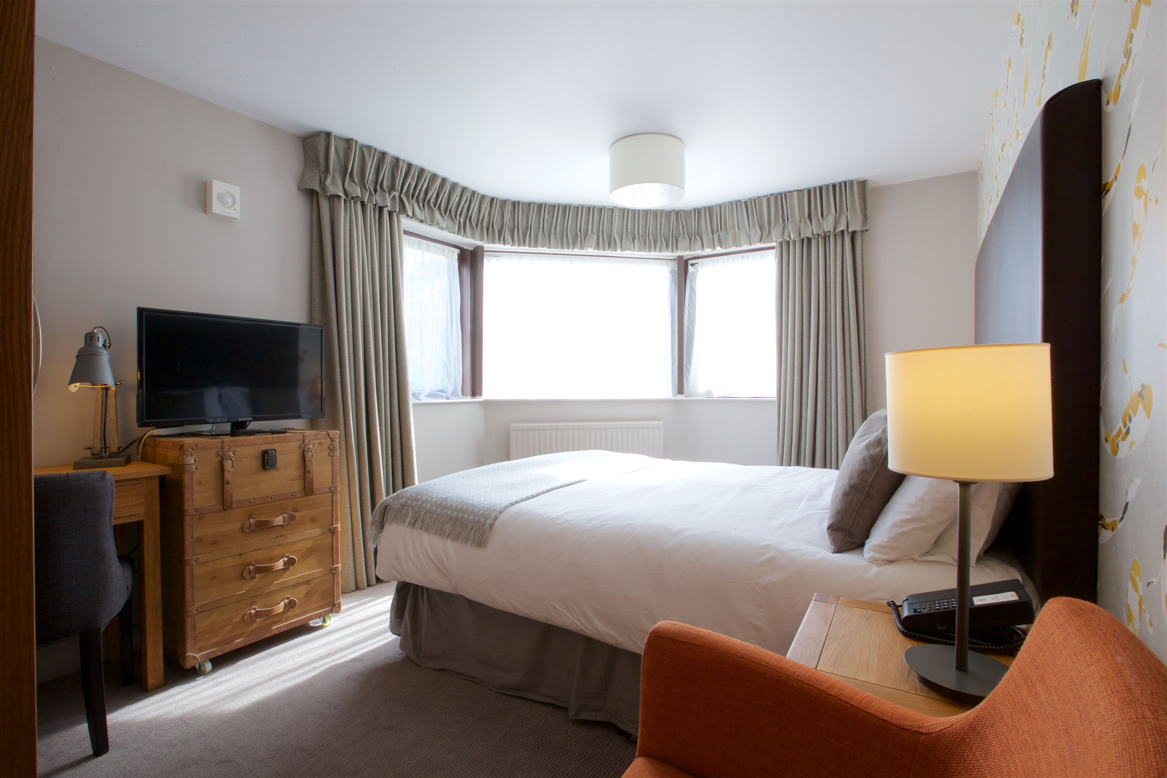 A hotel room with a bed, a desk with a television, a chair, and a lamp.