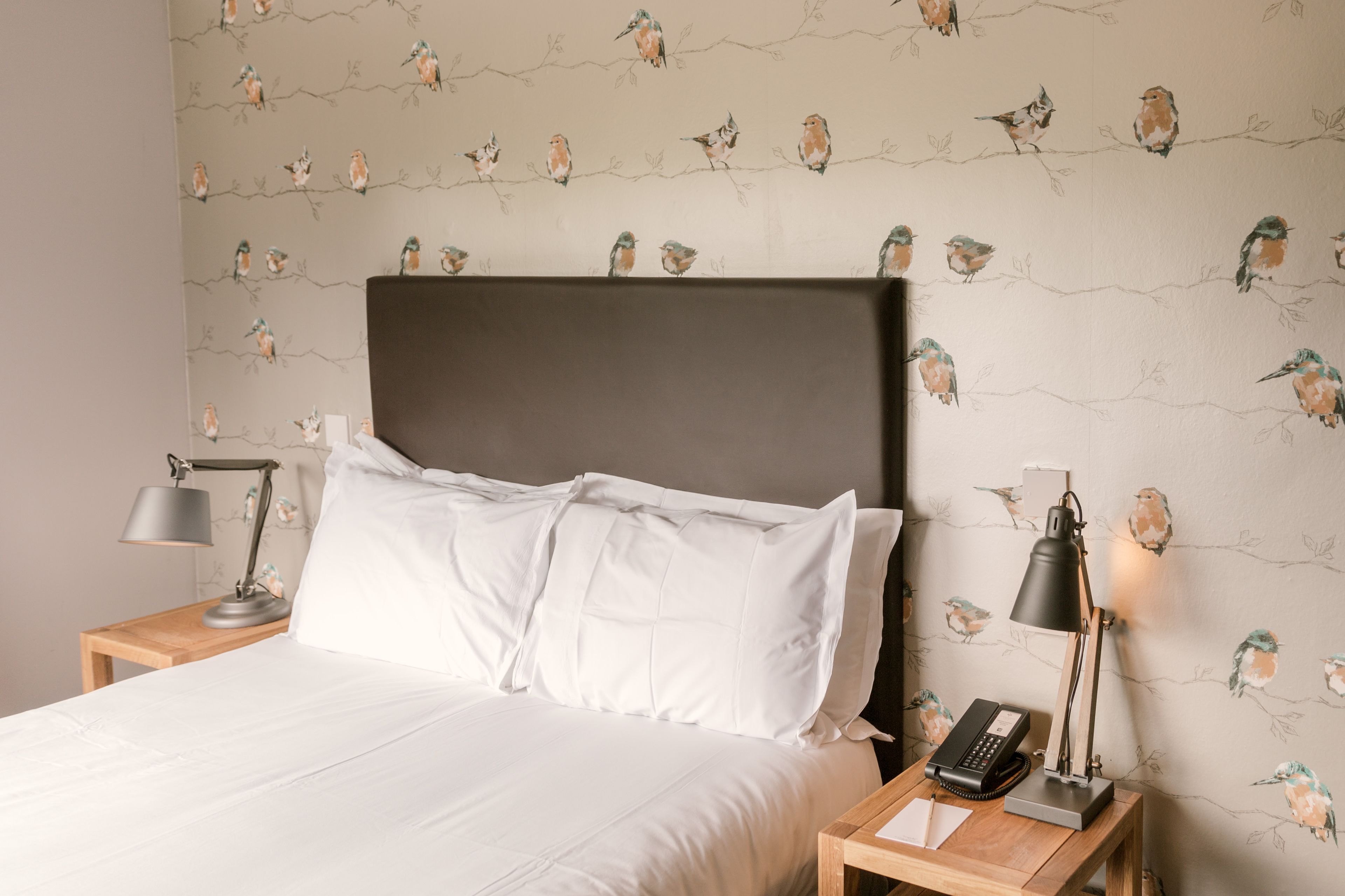 A neatly made bed with white linens, a dark headboard, and a bedside table with a lamp and a telephone.