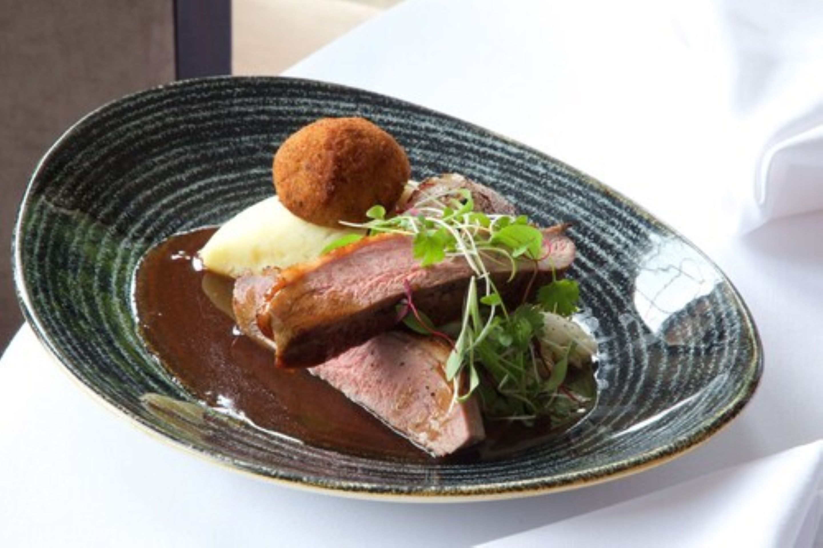 A plate with sliced meat, mashed potatoes, and a round item on a textured plate.