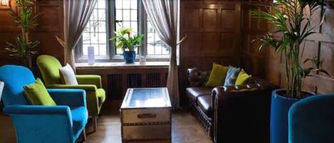 A cozy living room with a blue sofa, a brown leather couch, and a wooden coffee table. There are potted plants on the windowsill and a window with curtains.