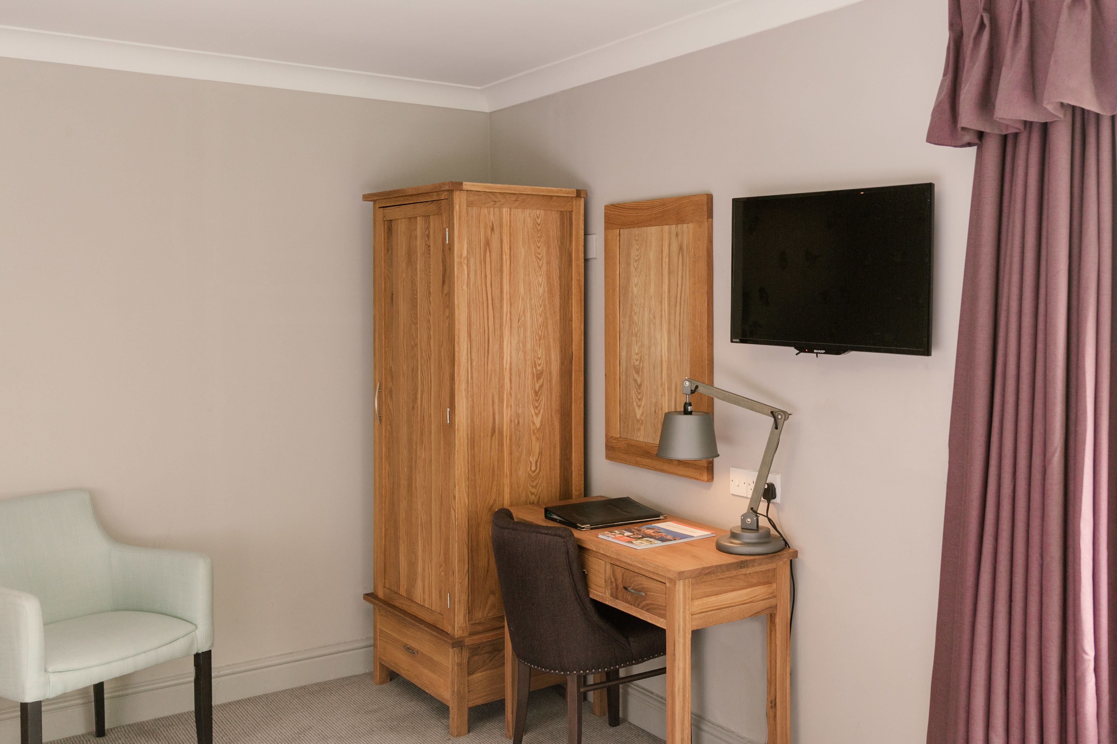A room with a wooden wardrobe, a desk with a chair, a flat-screen TV, and a desk lamp.