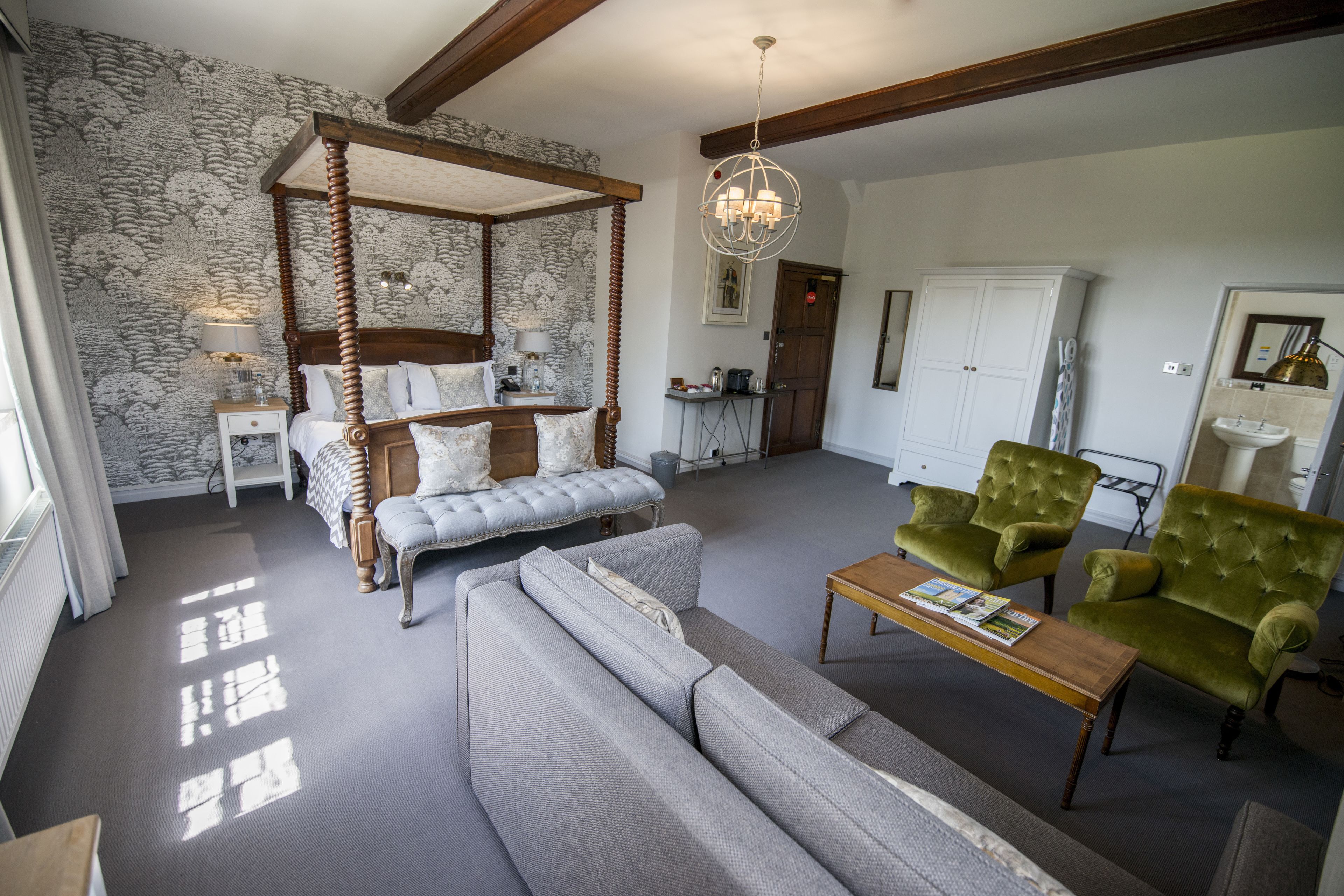 A four-poster bed with a wooden frame, a grey sofa, a green armchair, and a wooden coffee table in a well-lit room with patterned wallpaper and a chandelier.