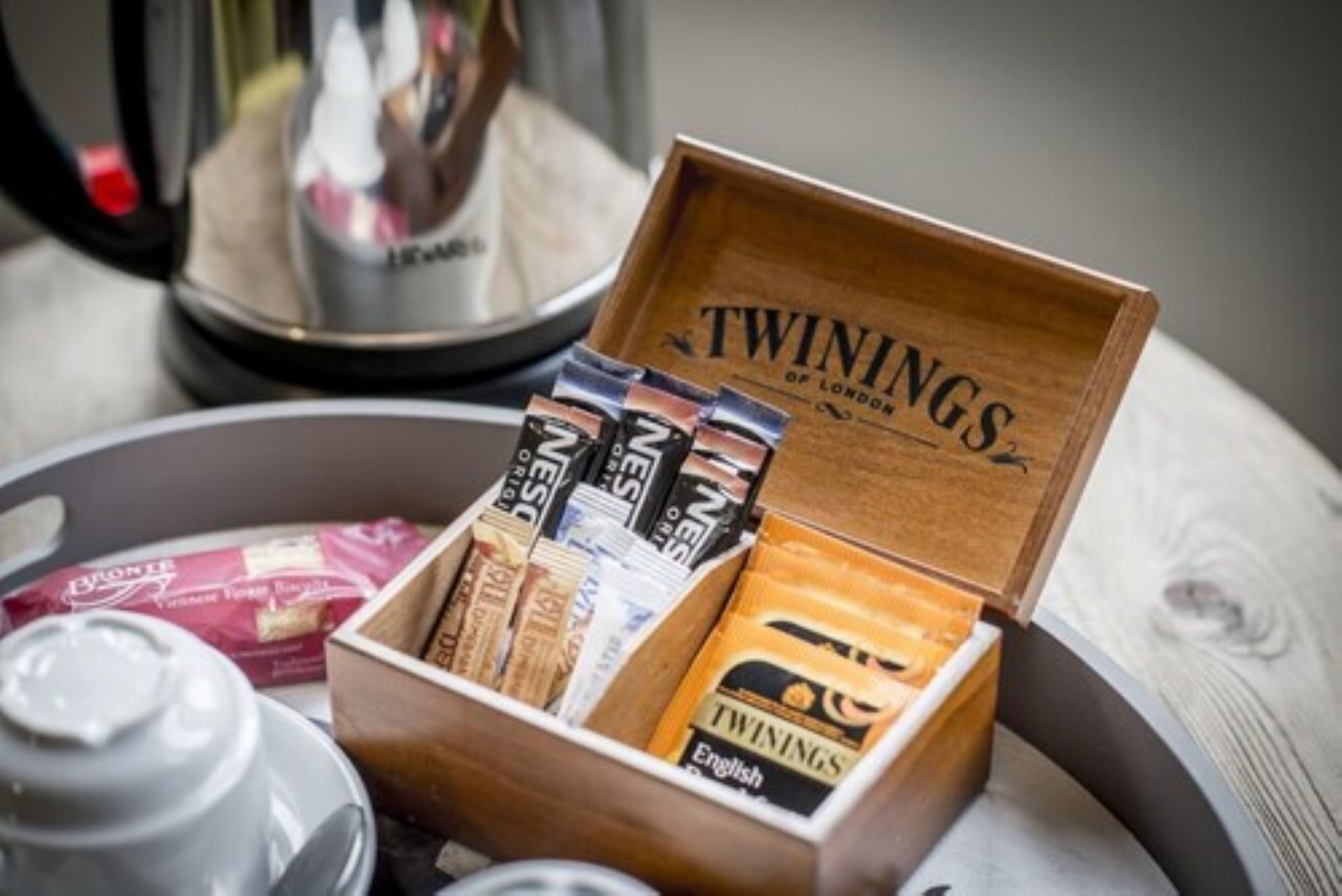 Una caja de madera con la etiqueta 'TWININGS OF LONDON' que contiene varios paquetes de té de la marca, una tetera y una taza sobre una bandeja.