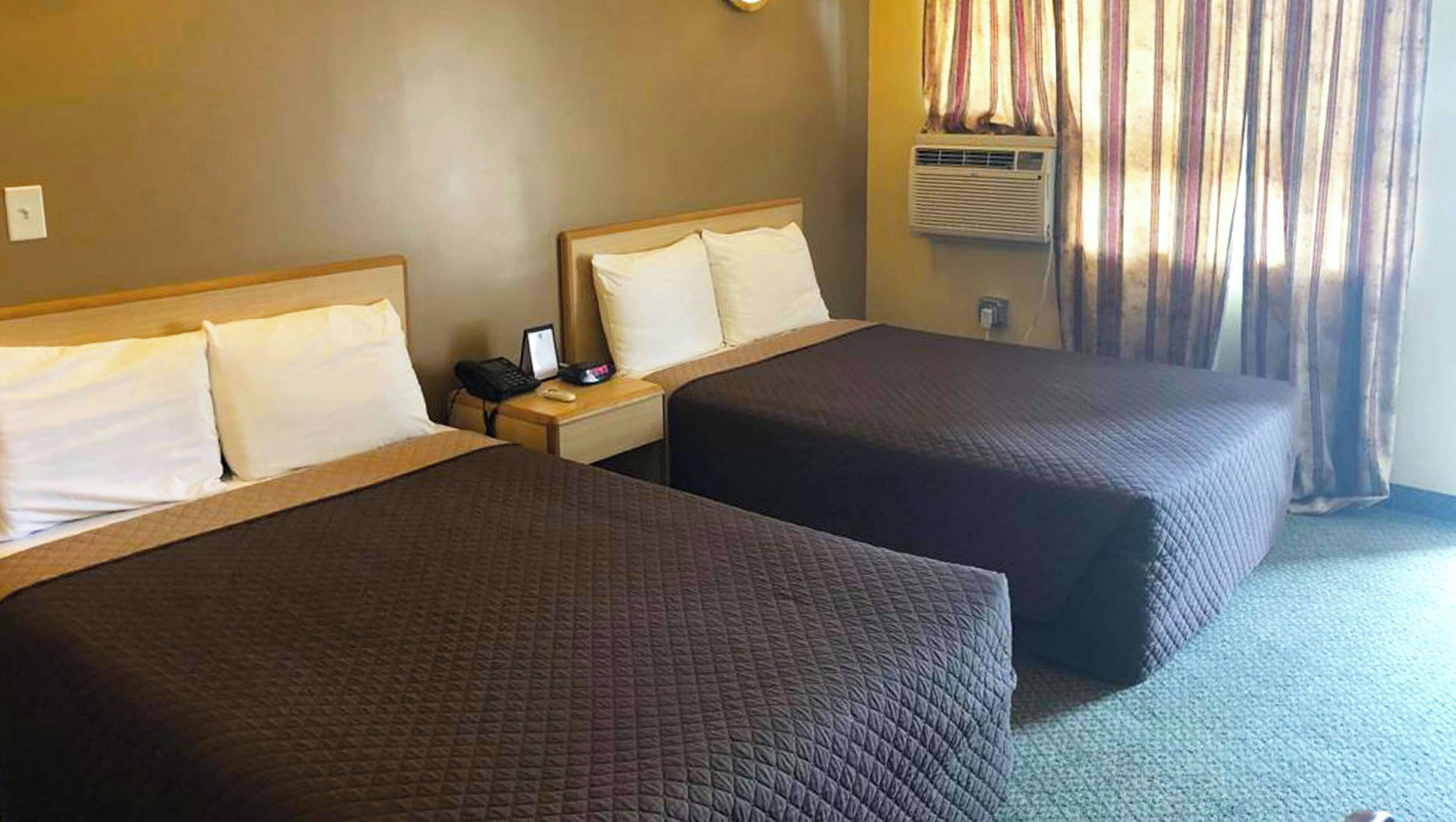 A hotel room with two beds, a nightstand with a phone and a lamp, and a window with curtains.