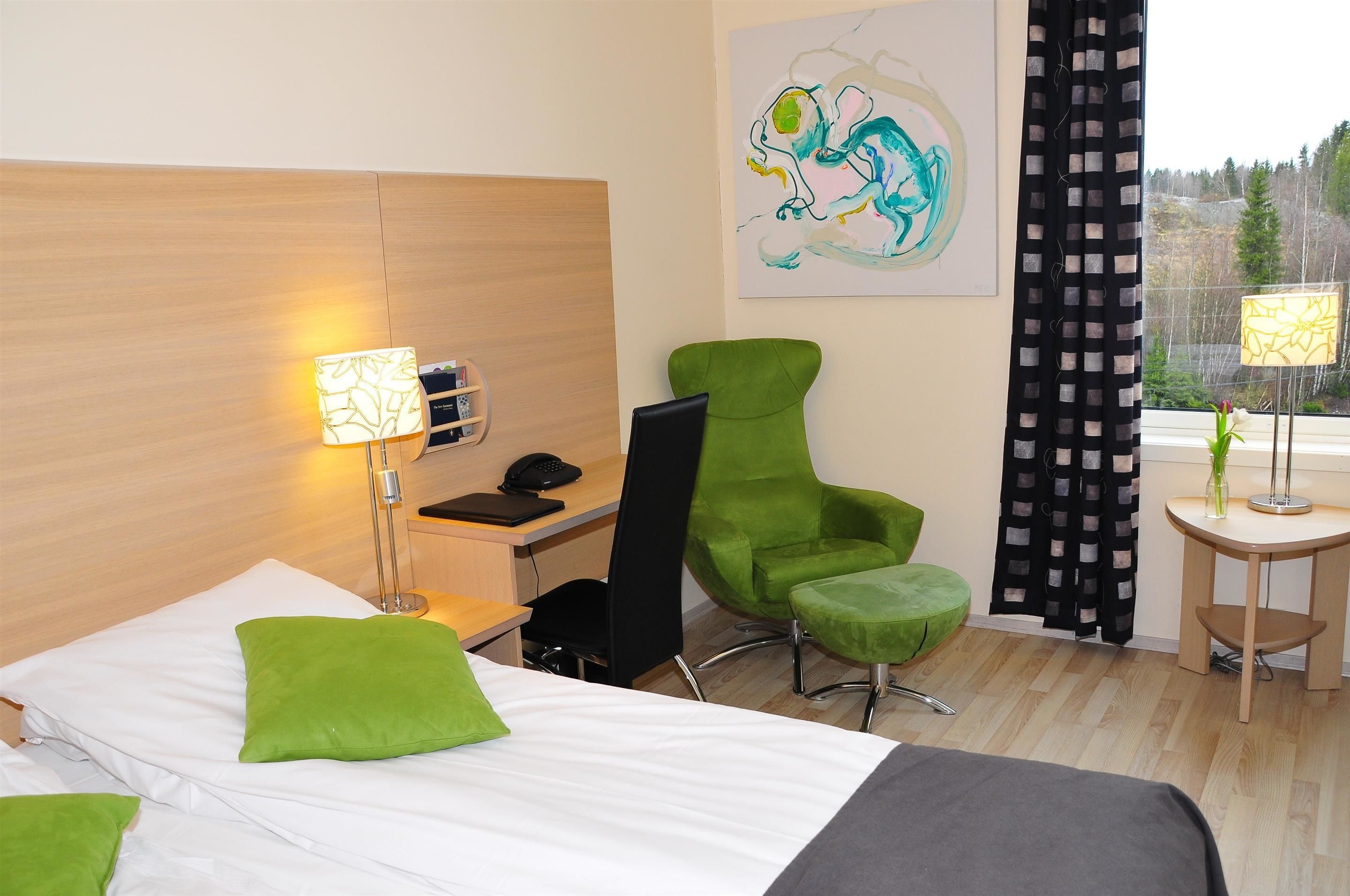 Een hotelkamer met een bed, groene kussens, een bureau met telefoon, een groene stoel, een klein tafeltje met lamp en een schilderij aan de muur.